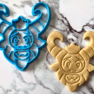 K&ouml;nnte beinhalten: Ein blauer Plastik-Ausstecher und ein gebackener Keks, beide in Form einer Comicfigur mit H&ouml;rnern, einer Krone und einem &uuml;berraschten Ausdruck.