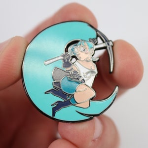 May include: A teal blue enamel pin featuring a cartoon character holding a pickaxe, sitting on a crescent moon.