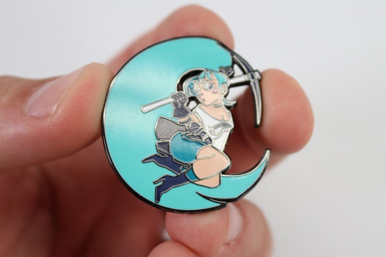 May include: A teal blue enamel pin featuring a cartoon character holding a pickaxe, sitting on a crescent moon.