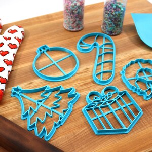 Op de afbeelding: Een set blauwe plastic kerstkoekjesvormen op een houten snijplank. De vormen zijn in de vorm van een kerstboom, een snoepstok, een cadeau, een ornament en een krans.