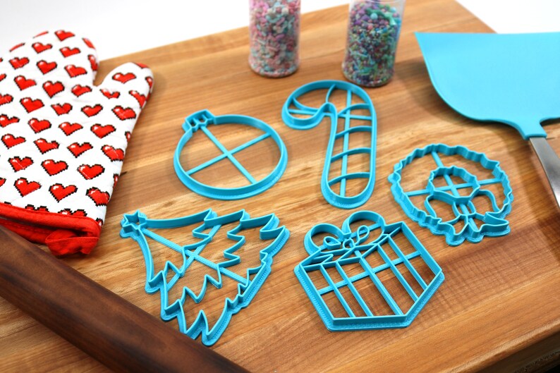 Op de afbeelding: Een set blauwe plastic kerstkoekjesvormen op een houten snijplank. De vormen zijn in de vorm van een kerstboom, een snoepstok, een cadeau, een ornament en een krans.