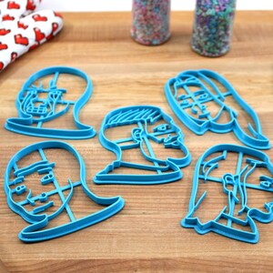 Op de afbeelding: Zes blauwe plastic koekjesvormen in de vorm van verschillende gezichten. De vormen liggen op een houten oppervlak.