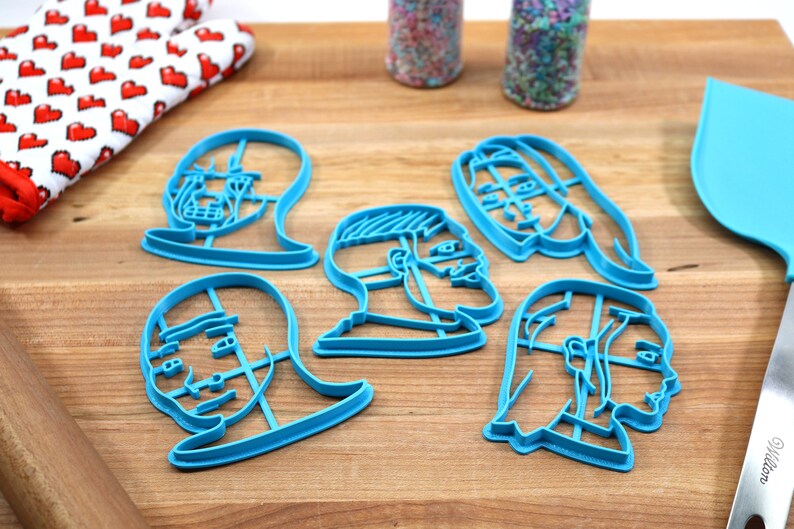 Op de afbeelding: Zes blauwe plastic koekjesvormen in de vorm van verschillende gezichten. De vormen liggen op een houten oppervlak.