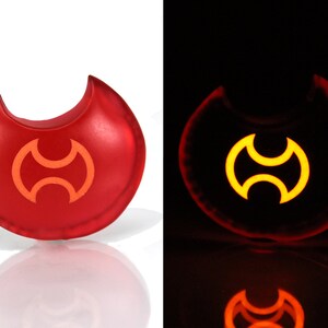 May include: A red glow-in-the-dark keychain shaped like a crescent moon with a stylized symbol in the center. The symbol is a double-headed axe with a circular design.
