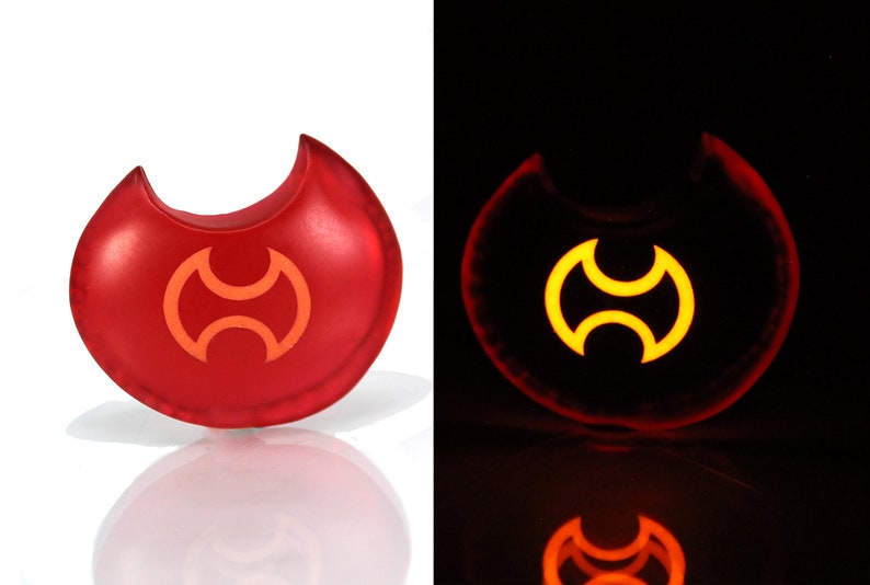 May include: A red glow-in-the-dark keychain shaped like a crescent moon with a stylized symbol in the center. The symbol is a double-headed axe with a circular design.