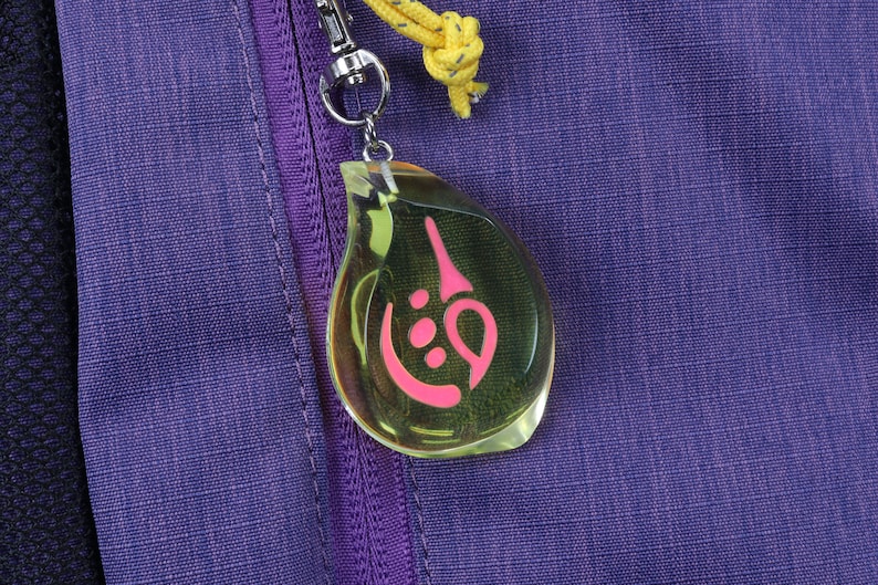 May include: A clear teardrop-shaped keychain with a pink symbol inside. The keychain is attached to a purple zipper on a bag.