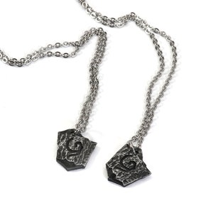 May include: Two silver necklaces with matching pendants. Each pendant is a silver shield shape with a textured surface and a stylized letter 'G' in the center.