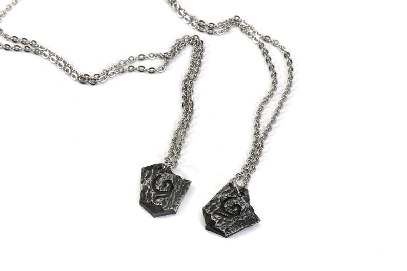 May include: Two silver necklaces with matching pendants. Each pendant is a silver shield shape with a textured surface and a stylized letter 'G' in the center.