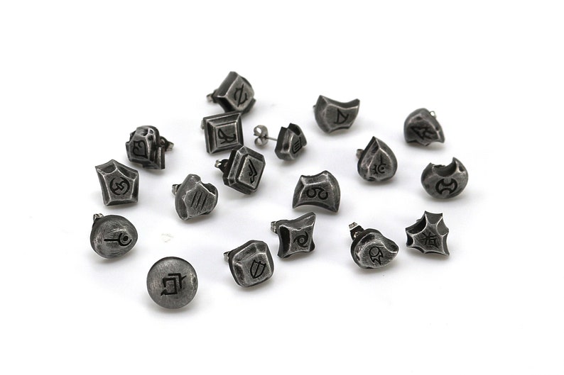 May include: A collection of 16 silver-toned earrings with various symbols and designs. The earrings are scattered on a white surface.
