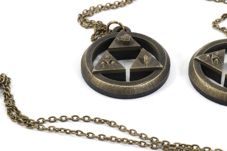 Puede incluir: Dos collares de color dorado con colgantes que representan el s&iacute;mbolo Triforce de la serie de videojuegos The Legend of Zelda. Cada colgante tiene un dise&ntilde;o circular con una estrella de tres puntas en el centro, con cada punta de la estrella representando un s&iacute;mbolo diferente.