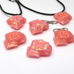 May include: Five pink resin keychains with a heart design. The keychains are made of a translucent pink resin with glitter embedded in the resin. The heart design is a raised, white heart with a pink outline. Two of the keychains are attached to black cords.