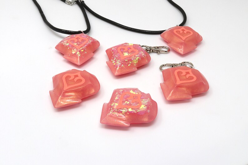 May include: Five pink resin keychains with a heart design. The keychains are made of a translucent pink resin with glitter embedded in the resin. The heart design is a raised, white heart with a pink outline. Two of the keychains are attached to black cords.