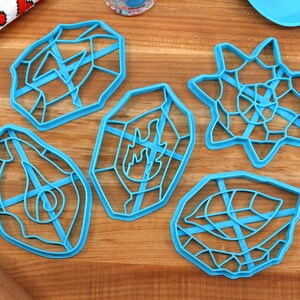 May include: Five blue plastic cookie cutters shaped like geometric designs. The cutters are on a wooden surface.
