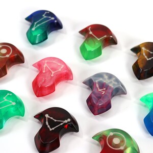 May include: Nine resin dice with different colours and constellations. The dice are shaped like a crescent moon with a pointed tip.