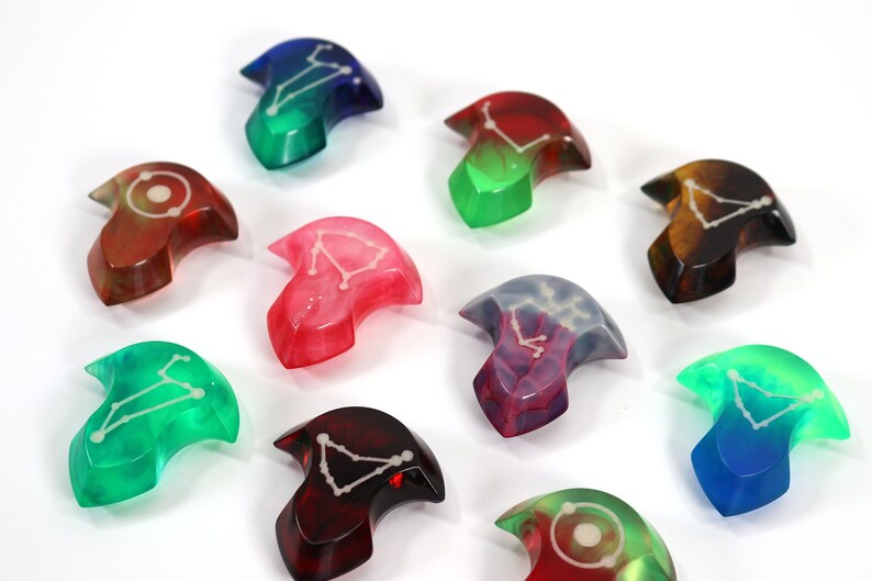 May include: Nine resin dice with different colours and constellations. The dice are shaped like a crescent moon with a pointed tip.