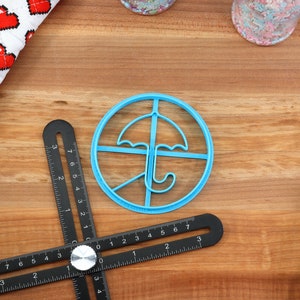 May include: A light blue plastic umbrella-shaped cookie cutter sits on a wooden surface, next to a black multi-angle measuring tool. A white and red heart-patterned cloth is in the upper left corner. Sprinkles and a blue spatula are also visible.