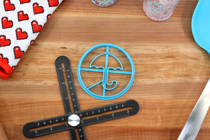 May include: A light blue plastic umbrella-shaped cookie cutter sits on a wooden surface, next to a black multi-angle measuring tool. A white and red heart-patterned cloth is in the upper left corner. Sprinkles and a blue spatula are also visible.