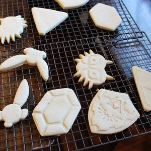 Puede incluir: Una rejilla de enfriamiento con nueve galletas de az&uacute;car simples decoradas con varios dise&ntilde;os, incluyendo un tri&aacute;ngulo, un hex&aacute;gono, un escudo, un conejo, un p&aacute;jaro y una cara de monstruo.