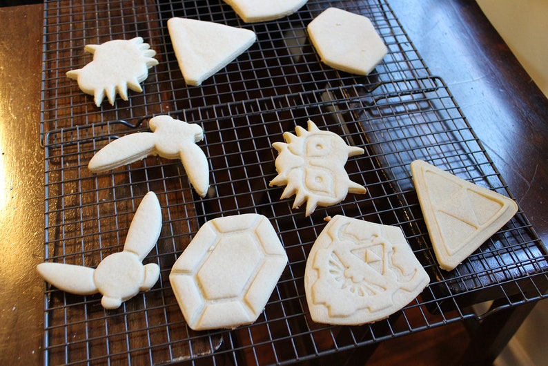 Puede incluir: Una rejilla de enfriamiento con nueve galletas de az&uacute;car simples decoradas con varios dise&ntilde;os, incluyendo un tri&aacute;ngulo, un hex&aacute;gono, un escudo, un conejo, un p&aacute;jaro y una cara de monstruo.