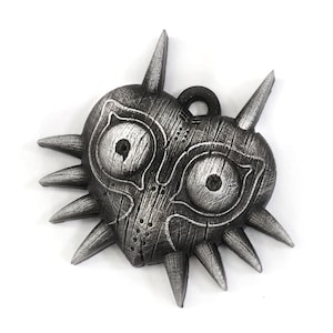 May include: A silver-colored Majora's Mask pendant with a heart shape and sharp spikes around the edges. The mask has two large eyes and a detailed wooden texture.