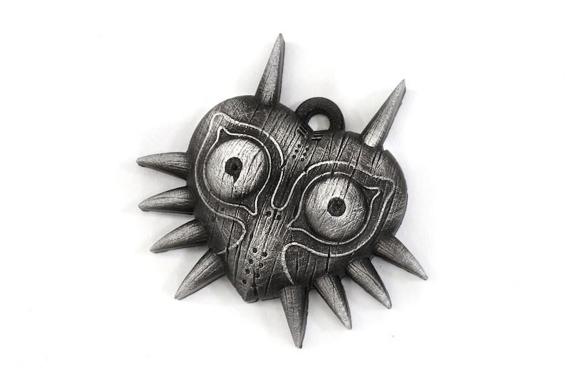 May include: A silver-colored Majora's Mask pendant with a heart shape and sharp spikes around the edges. The mask has two large eyes and a detailed wooden texture.