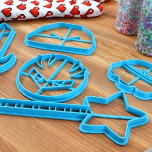May include: A set of six blue plastic cookie cutters shaped like a sword, a star, a wand, a cupcake, a helmet, and a shield. The cutters are on a wooden surface.