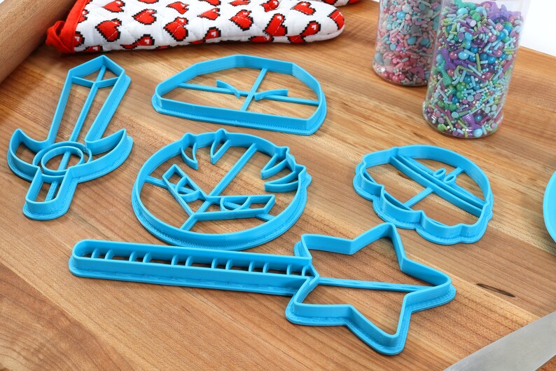 May include: A set of six blue plastic cookie cutters shaped like a sword, a star, a wand, a cupcake, a helmet, and a shield. The cutters are on a wooden surface.