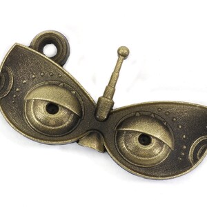 May include: A gold-colored metal charm with a whimsical design featuring two large eyes and a small antenna. The charm has a textured surface and a loop for attaching to a necklace or bracelet.