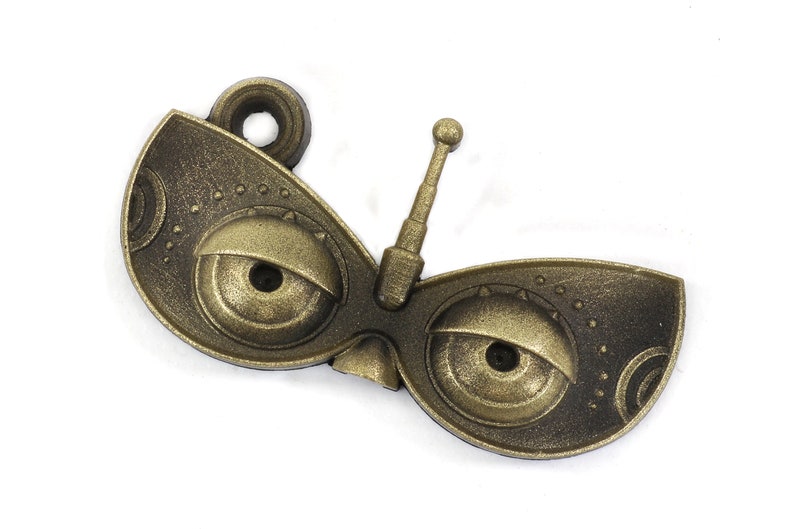 May include: A gold-colored metal charm with a whimsical design featuring two large eyes and a small antenna. The charm has a textured surface and a loop for attaching to a necklace or bracelet.