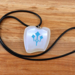 May include: A white pendant with a blue symbol on a black cord necklace. The pendant is shaped like a shield and has a smooth, glossy finish.
