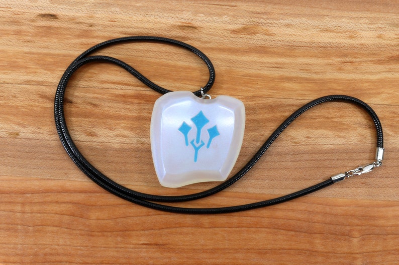 May include: A white pendant with a blue symbol on a black cord necklace. The pendant is shaped like a shield and has a smooth, glossy finish.
