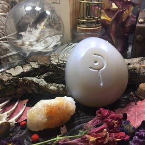 May include: A white egg-shaped stone with a carved symbol on it. The stone is surrounded by various objects, including a clear glass orb, a piece of driftwood, and a small glass jar with a cork stopper.