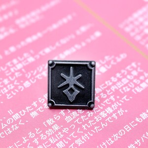 May include: A square, silver-colored pin with a stylized symbol in the center. The pin is set against a pink background with Japanese text.