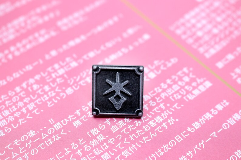 May include: A square, silver-colored pin with a stylized symbol in the center. The pin is set against a pink background with Japanese text.