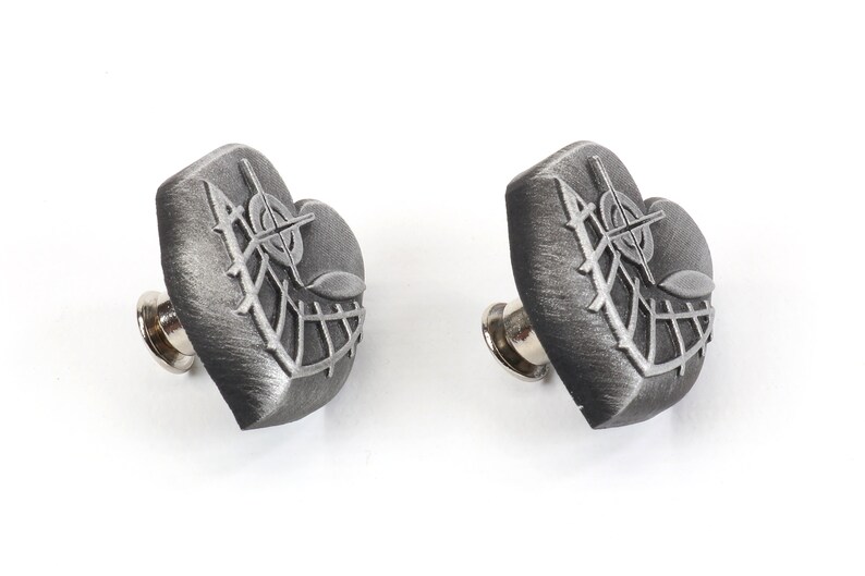 K&ouml;nnte beinhalten: Zwei silberne herzf&ouml;rmige Anstecknadeln mit einem detaillierten Design aus einem Kreuz und Bl&auml;ttern. Die Anstecknadeln haben eine silberne Metallnadelr&uuml;ckseite.