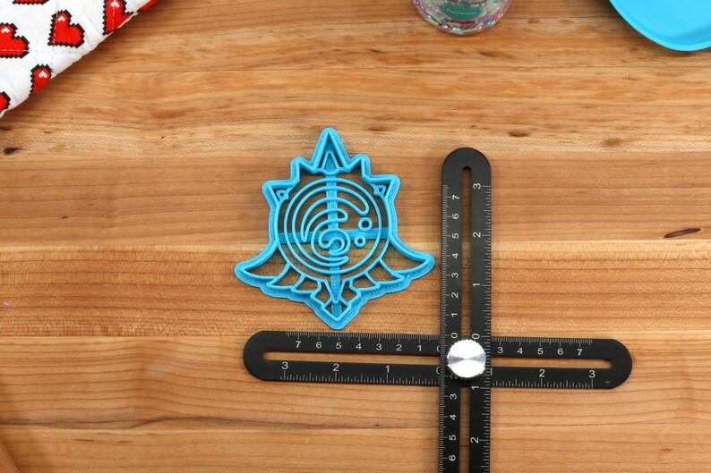 May include: A blue plastic cookie cutter shaped like a stylized sun with a swirling design inside. The cutter is about 2 inches in diameter and is sitting on a wooden surface.