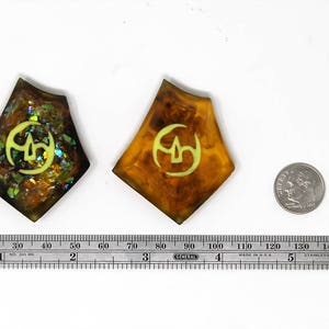 May include: Two resin pendants with a gold and green iridescent finish. Each pendant features a yellow symbol on a brown background. A ruler and a US quarter are included for scale.