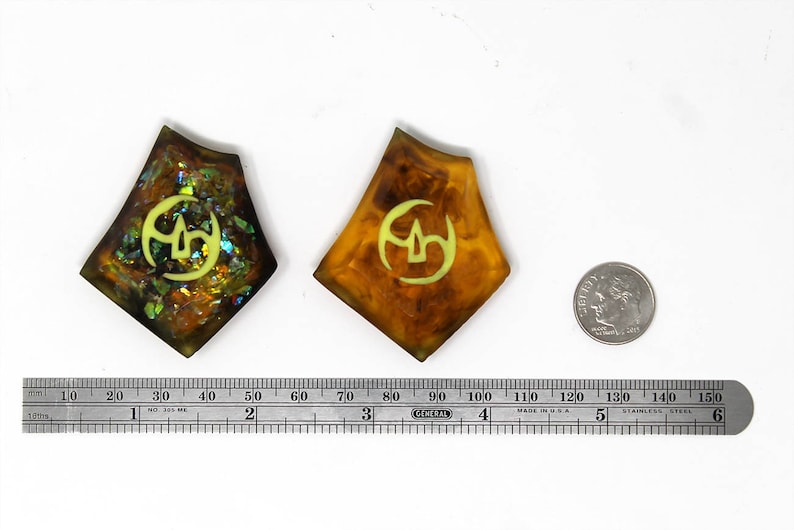 May include: Two resin pendants with a gold and green iridescent finish. Each pendant features a yellow symbol on a brown background. A ruler and a US quarter are included for scale.
