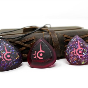 May include: Three teardrop-shaped pendants with a pink symbol design. The pendants are purple and red with iridescent glitter. A black cord is attached to one pendant. A brown leather journal is in the background.