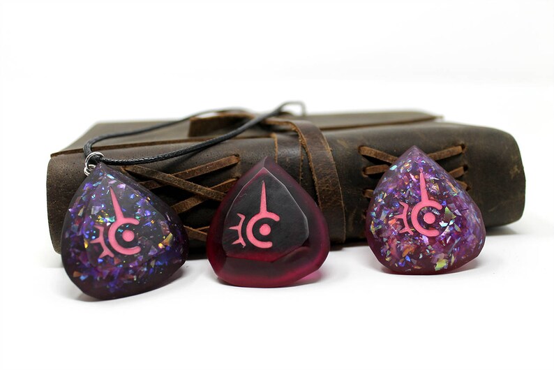 May include: Three teardrop-shaped pendants with a pink symbol design. The pendants are purple and red with iridescent glitter. A black cord is attached to one pendant. A brown leather journal is in the background.