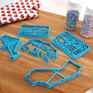 May include: A set of six blue plastic cookie cutters shaped like a California bear, a California outline, a palm tree, a Golden Gate Bridge, and two different California landscapes. The cutters are on a wooden surface with a rolling pin, a pair of oven mitts, and two jars of sprinkles.