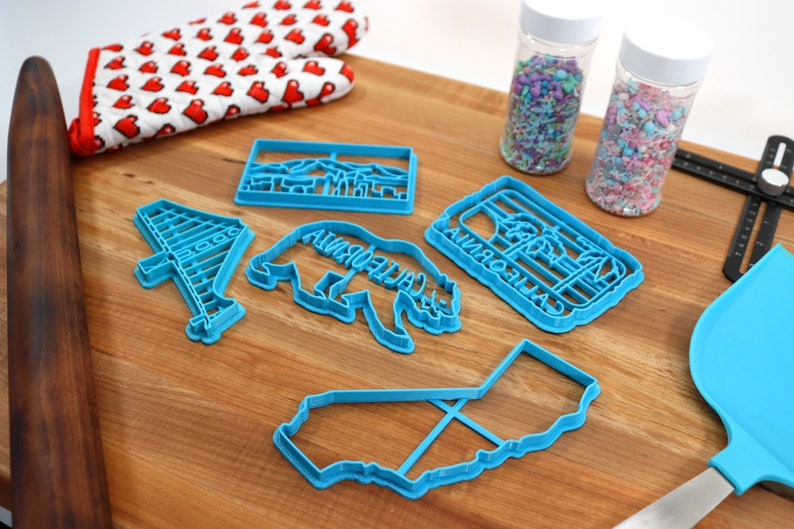 May include: A set of six blue plastic cookie cutters shaped like a California bear, a California outline, a palm tree, a Golden Gate Bridge, and two different California landscapes. The cutters are on a wooden surface with a rolling pin, a pair of oven mitts, and two jars of sprinkles.