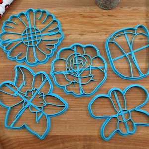 May include: Set of five blue plastic cookie cutters shaped like different flowers. The cutters are on a wooden surface.