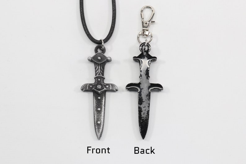 May include: A silver dagger pendant with intricate details on the hilt, shown on a black cord necklace and as a keychain. The dagger has a distressed, aged look.