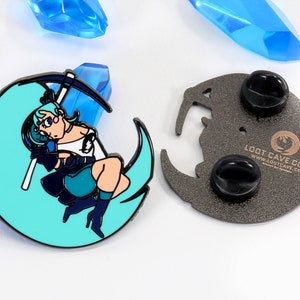 May include: A blue and black enamel pin featuring a cartoon character holding a pickaxe, standing on a crescent moon. The back of the pin is silver with two black pin backs and the text "Loot Cave Co. www.lootcave.com".