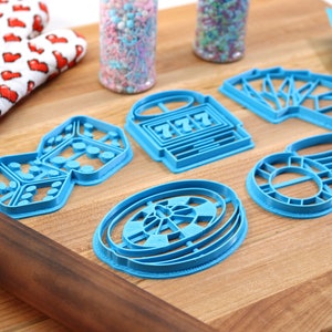 May include: A set of six blue plastic cookie cutters shaped like dice, a slot machine, playing cards, a roulette wheel, and a stack of chips. The cutters are on a wooden surface.