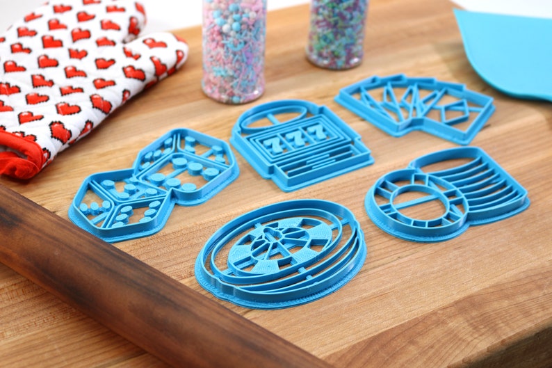 May include: A set of six blue plastic cookie cutters shaped like dice, a slot machine, playing cards, a roulette wheel, and a stack of chips. The cutters are on a wooden surface.