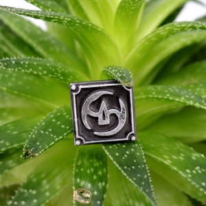May include: A silver-colored metal pin with a geometric design. The pin is on a green plant with small white dots.