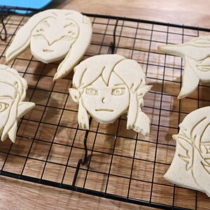 May include: Five sugar cookies decorated with icing in the shape of cartoon faces. The cookies are on a black wire cooling rack.