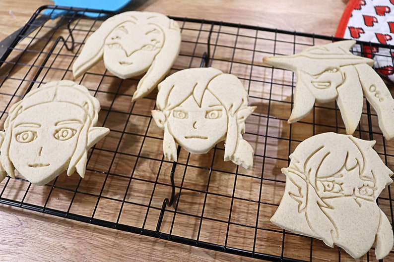 May include: Five sugar cookies decorated with icing in the shape of cartoon faces. The cookies are on a black wire cooling rack.